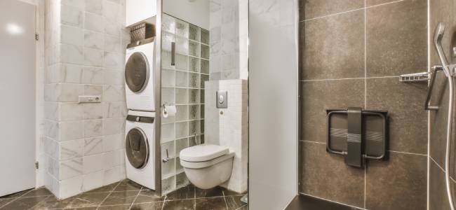 A bathroom with a wall-mounted toilet, glass shower, and stacked washer and dryer. The walls are tiled and the floor has dark tiles.