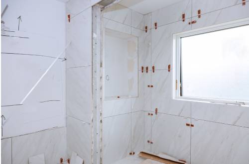 A bathroom under renovation with white marble tiles partially installed on the walls, and a window on the right.