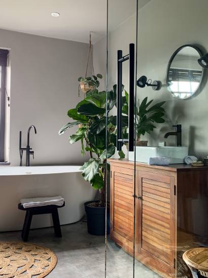 Modern bathroom with a wooden vanity, round mirror, and black fixtures. A large potted plant and a bathtub with a black faucet are beside a window.