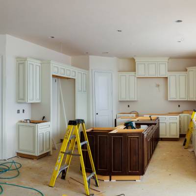 Kitchen under renovation with white cabinets, a wooden island, and multiple ladders. Walls are unpainted and floors uncovered. Tools and construction materials are visible.