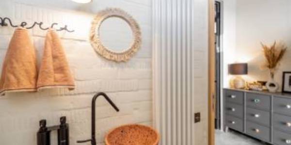 Bathroom with a round mirror, two towels on hooks, a black faucet over a textured sink, and a view into a room with a dresser and lamp.