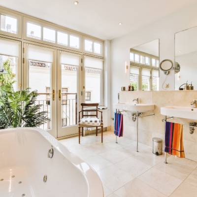 Spacious bathroom with large bathtub, two sinks, wall mirrors, and a cushioned chair. There is a potted plant and towels are hung beside the sinks. Natural light streams through the glass doors.