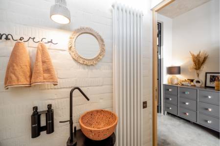 A bathroom with a round mirror, two orange towels, a black faucet with a brown vessel sink, and soap dispensers. A doorway leads to a room with a dresser and lamp.