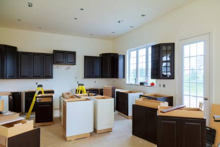 A kitchen under renovation with incomplete cabinetry, dark wood upper cabinets, and white lower cabinets. A ladder and some tools are visible, and large windows provide natural light.