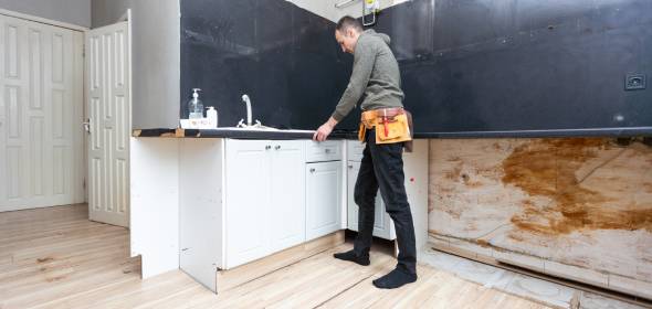 A person wearing a tool belt inspects the countertop in a partially renovated kitchen with white cabinets and black walls.