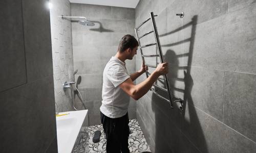 A man installs a towel rack in a modern bathroom with gray tile walls.