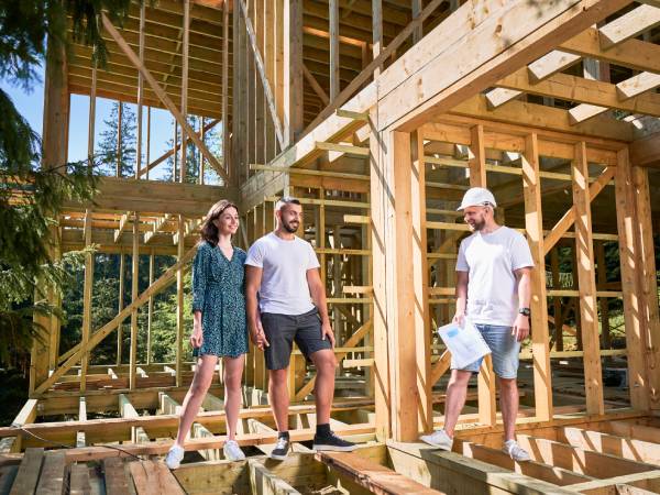 Three people stand on the wooden frame of a house under construction, surrounded by trees. One holds blueprints.